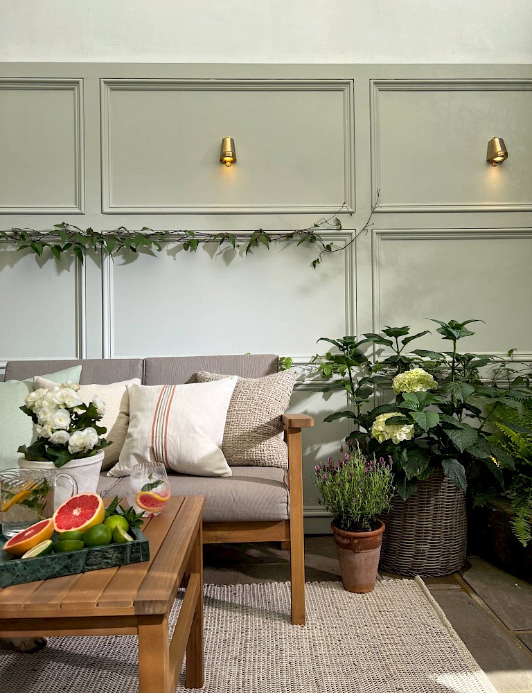 Kemptown courtyard garden design Brighton — sage green panelled walls, brass wall lights, teak outdoor sofa, woven rug and lush planting by Hestia & Hearth